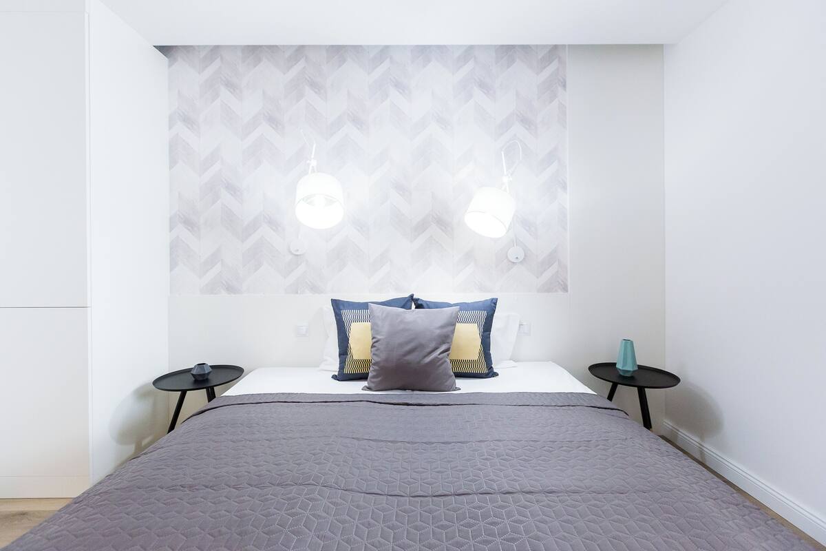 Ein modernes Schlafzimmer mit einem Kopfende im geometrischen Muster, einer grauen gesteppten Bettdecke, zwei Nachttischen mit Lampen und einer kleinen, türkisfarbenen Vase auf einem Tisch.