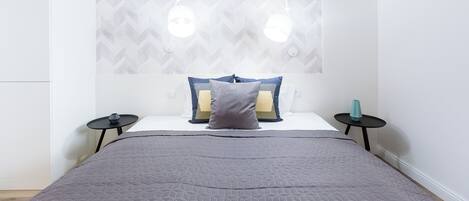 A modern bedroom with a geometric patterned headboard, a grey quilted bedspread, two bedside tables with lamps, and a small teal vase on one table.