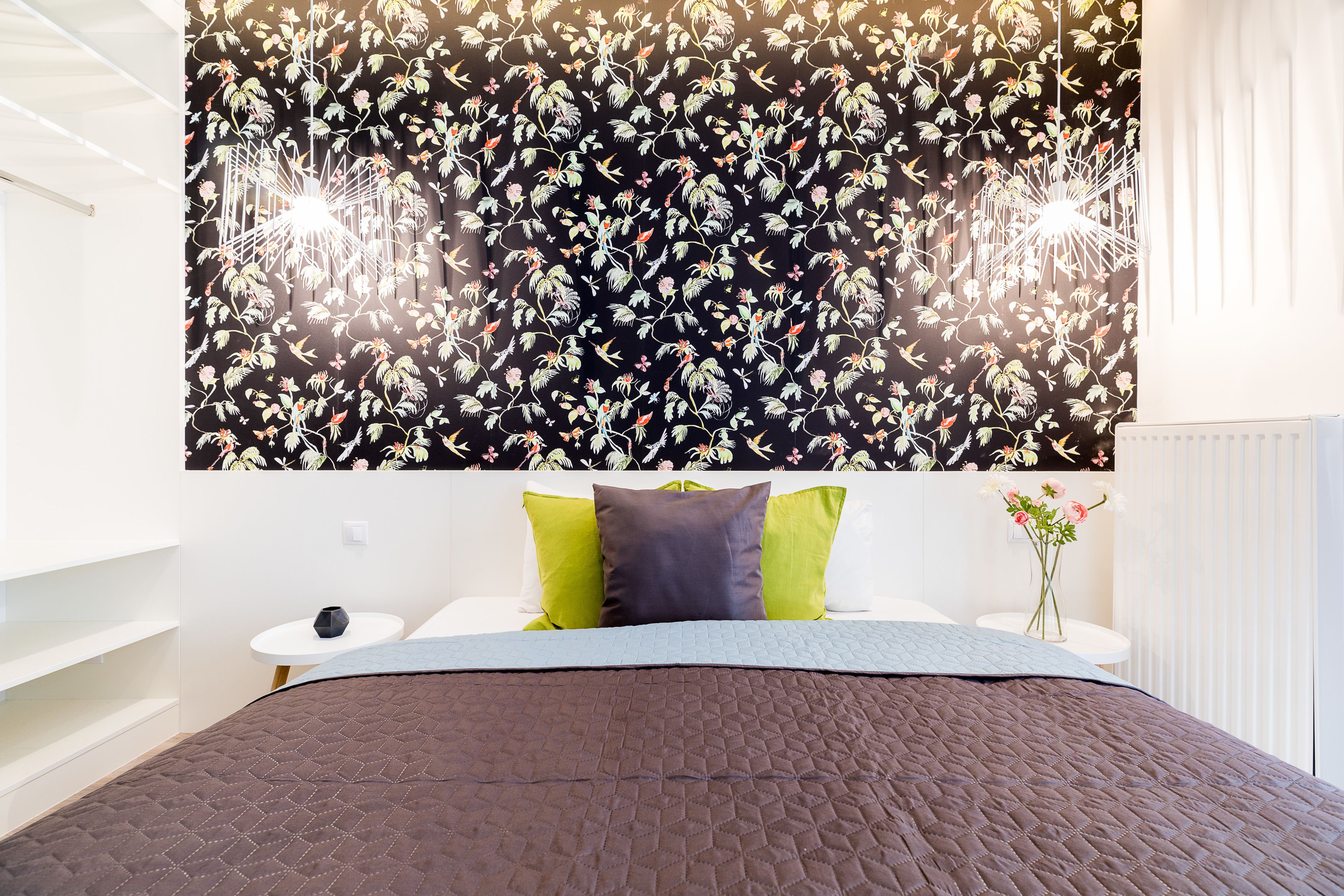 Une chambre avec un papier peint à motifs floraux, un lit recouvert d’une couette marron, deux coussins verts, une table de chevet blanche et un petit vase avec des fleurs.