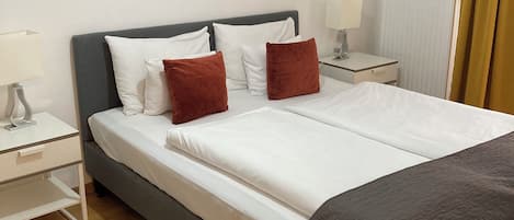 Ein ordentlich bezogenes Doppelbett mit weißer und brauner Bettwäsche, zwei Nachttische mit Lampen sowie ein Holzboden.