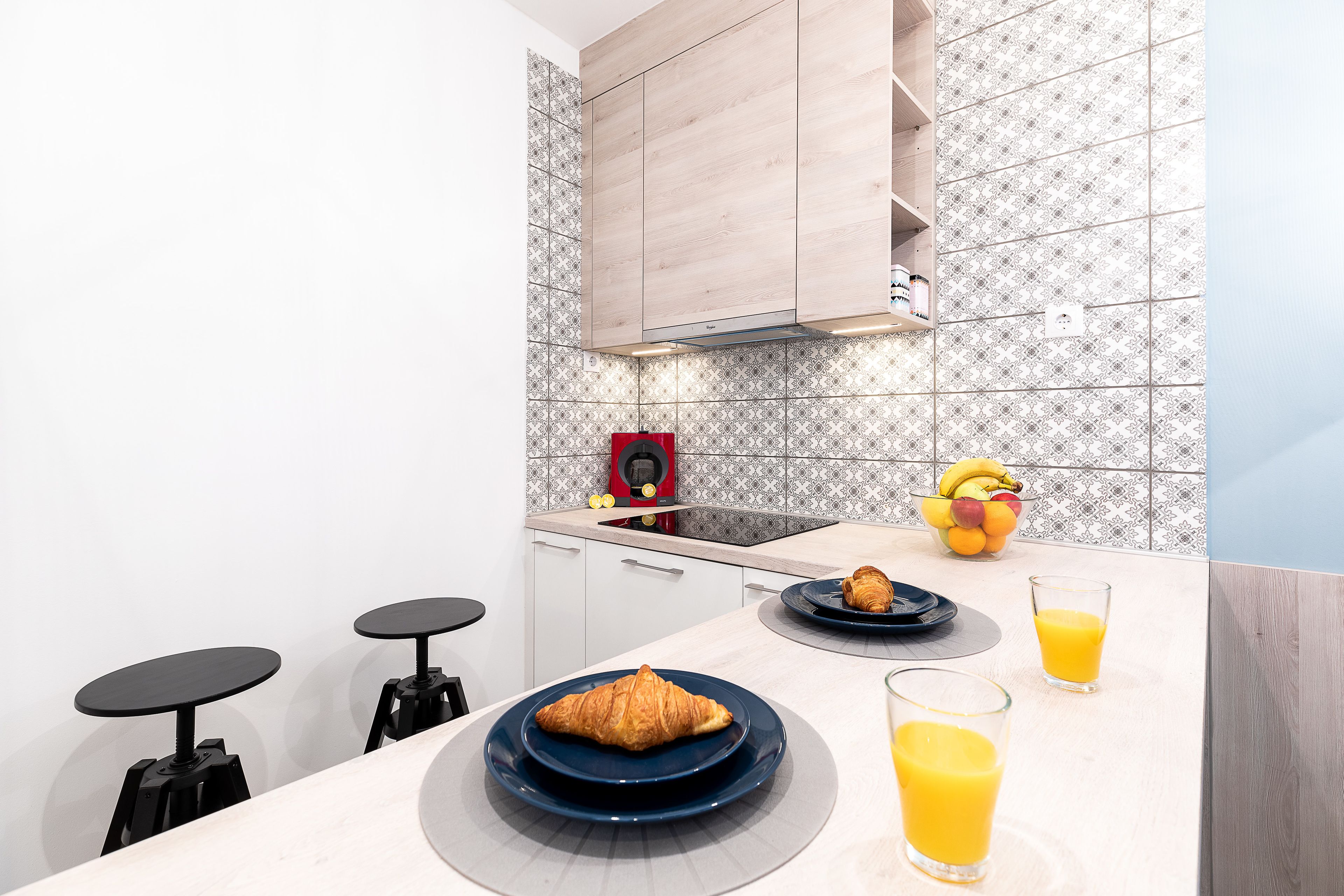 A modern kitchen with white cabinets, a patterned backsplash, and a breakfast setup including croissants, fruit, and orange juice.