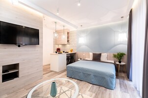 Ένα σύγχρονο δωμάτιο ξενοδοχείου με τηλεόραση επίπεδης οθόνης τοποθετημένη στον τοίχο, ένα κρεβάτι με μπλε κουβέρτα, μια μικρή κουζίνα και ένα τραπέζι φαγητού με καρέκλες.