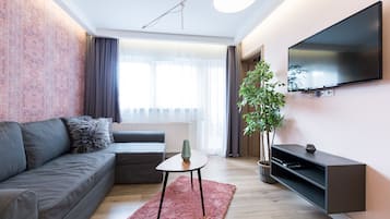 Ein modernes Wohnzimmer mit grauem Sofa, einem kleinen Tisch, einem an der Wand montierten Fernseher und einer Topfpflanze.