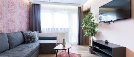A modern living room with a grey sofa, a small table, a TV mounted on the wall, and a potted plant.