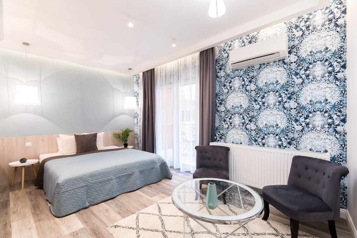 Ein modernes Schlafzimmer mit einem Bett, einem runden Glastisch, einem grauen Sessel und einem großen Fenster mit Vorhängen.
