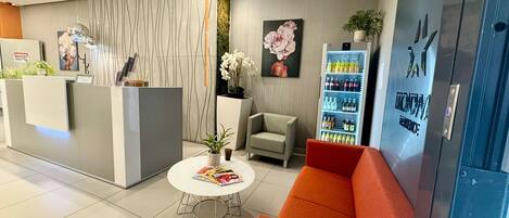 Zona de recepción con un mostrador moderno, un área de descanso con un sofá rojo y una pequeña mesa redonda, y un refrigerador azul lleno de bebidas.