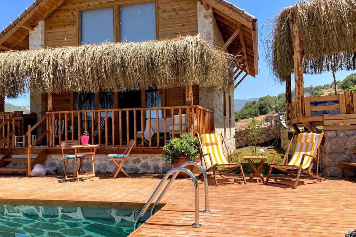 A wooden cabin with a thatched roof, a wooden deck, a pool, and lounge chairs.