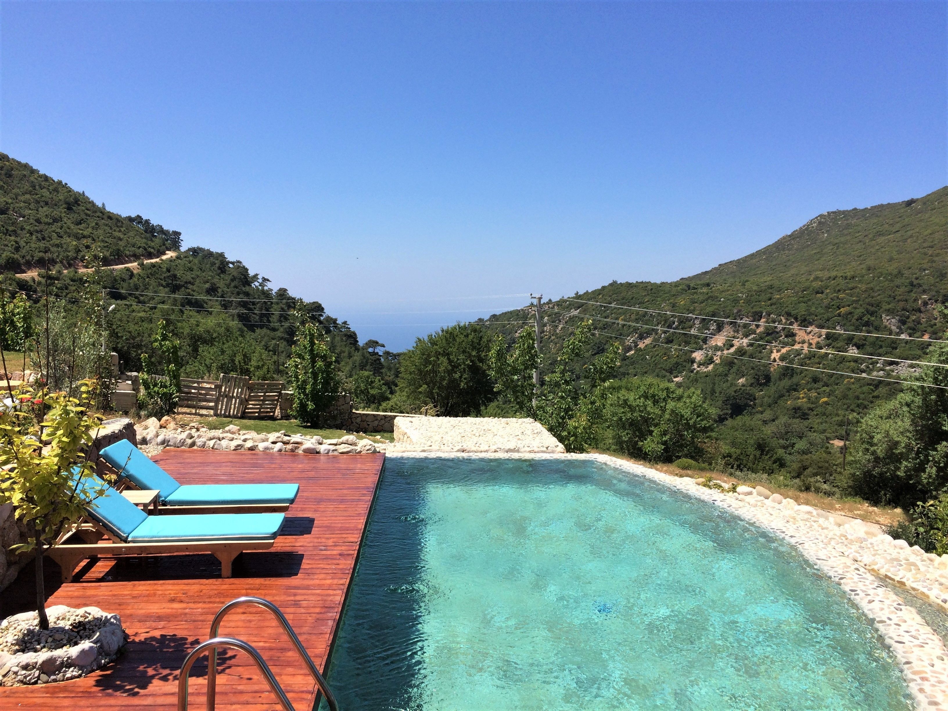 A pool with a wooden deck and lounge chairs, surrounded by lush greenery and hills.