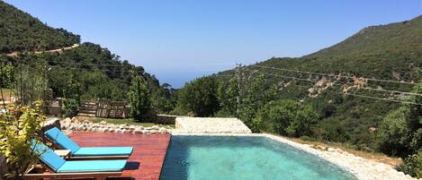 A pool with a wooden deck and lounge chairs, surrounded by lush greenery and hills.