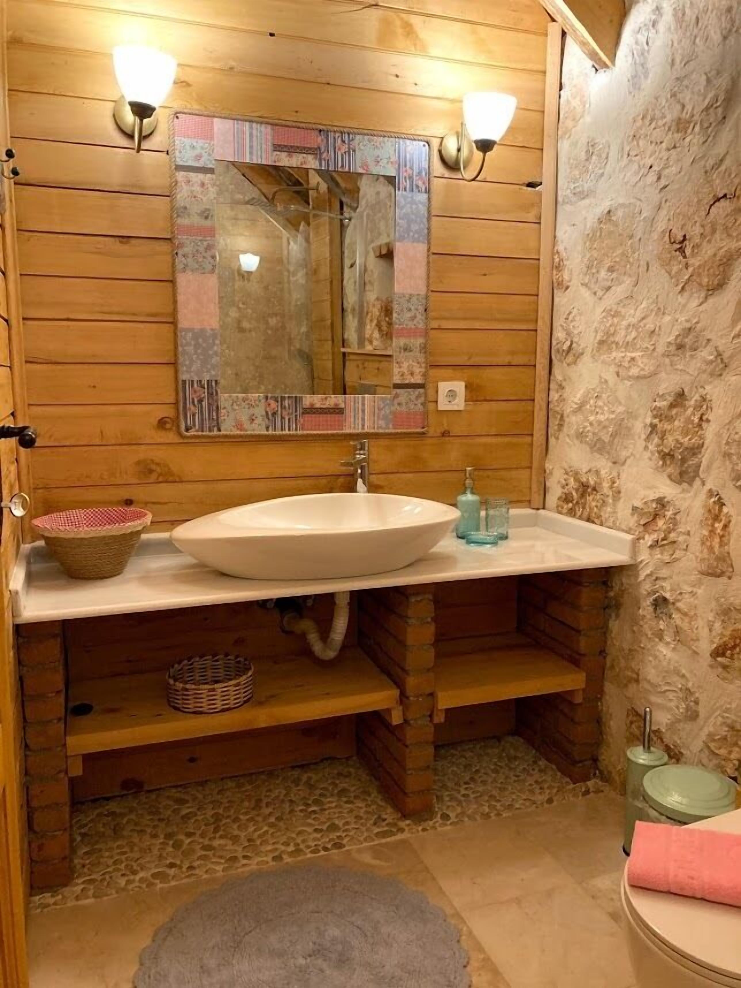 Un baño de madera con un espejo grande, dos luces empotradas en la pared, un lavamanos blanco y una pared de piedra.