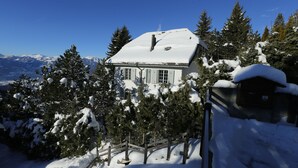 Une maison recouverte de neige, avec un escalier en bois menant à l’entrée, entourée d’arbres chargés de neige et avec des montagnes enneigées en arrière-plan.