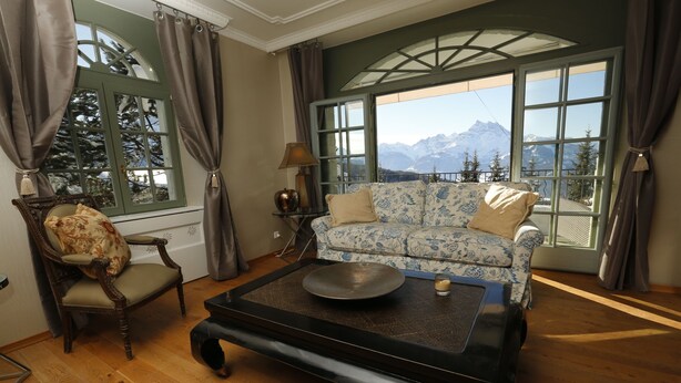 Une pièce avec un plancher en bois, un canapé recouvert d’un tissu à motifs, une table ronde et une chaise. De grandes fenêtres, ornées de rideaux, offrent une vue sur les montagnes.