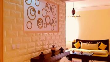 Une chambre d’hôtel moderne avec un mur en briques blanches, une œuvre d’art murale circulaire décorative, un canapé noir avec des coussins jaunes et une table ronde sur laquelle est posé un vase.