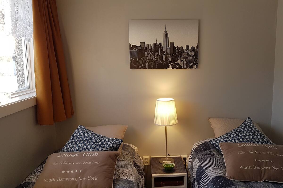 A hotel room with two beds, a cityscape wall art, a lamp, and a pillow with "Lounge Club The Harbour & Boathouse South Hamptons, New York" text.