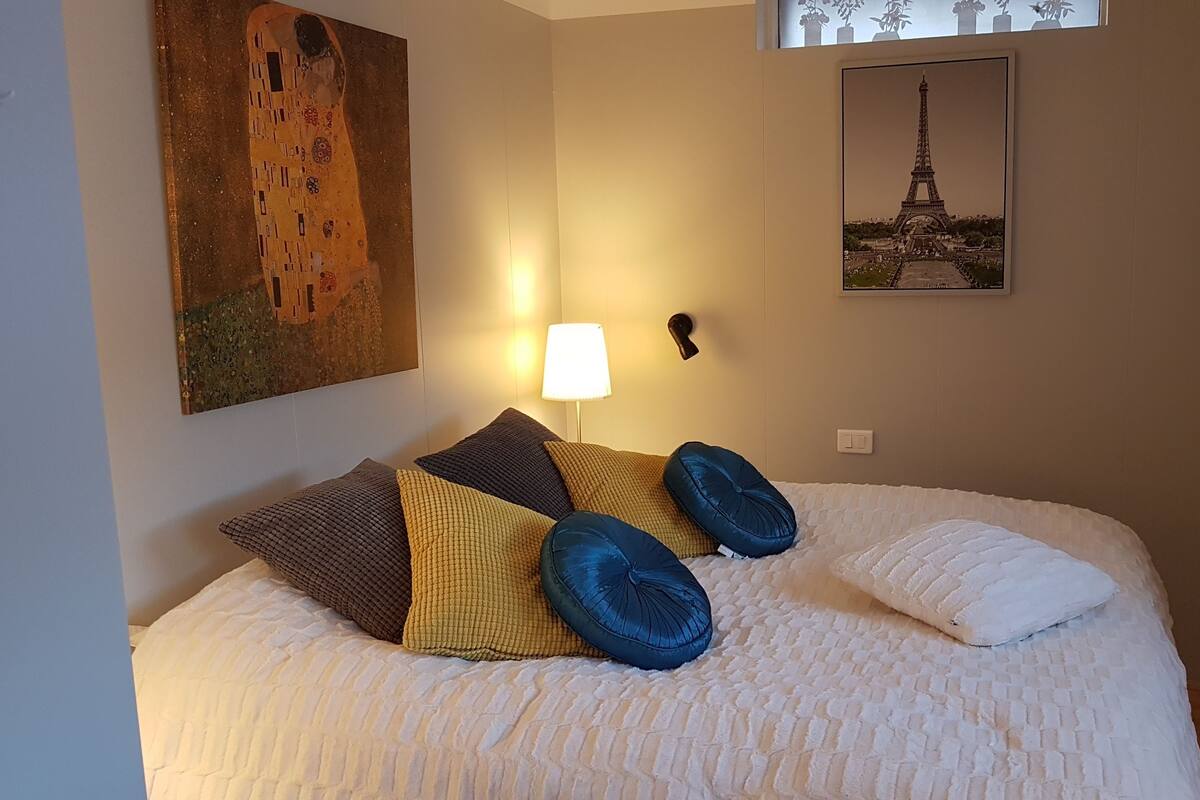 A neatly made bed with a white duvet, multiple pillows in various colors, a painting of a couple on the wall, and a framed picture of the Eiffel Tower.