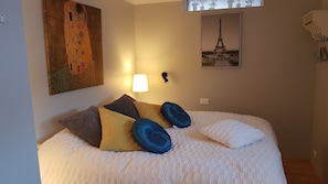 A neatly made bed with a white duvet, multiple pillows in various colors, a painting of a couple on the wall, and a framed picture of the Eiffel Tower. - The Viking Country Club (Hjalteyri)