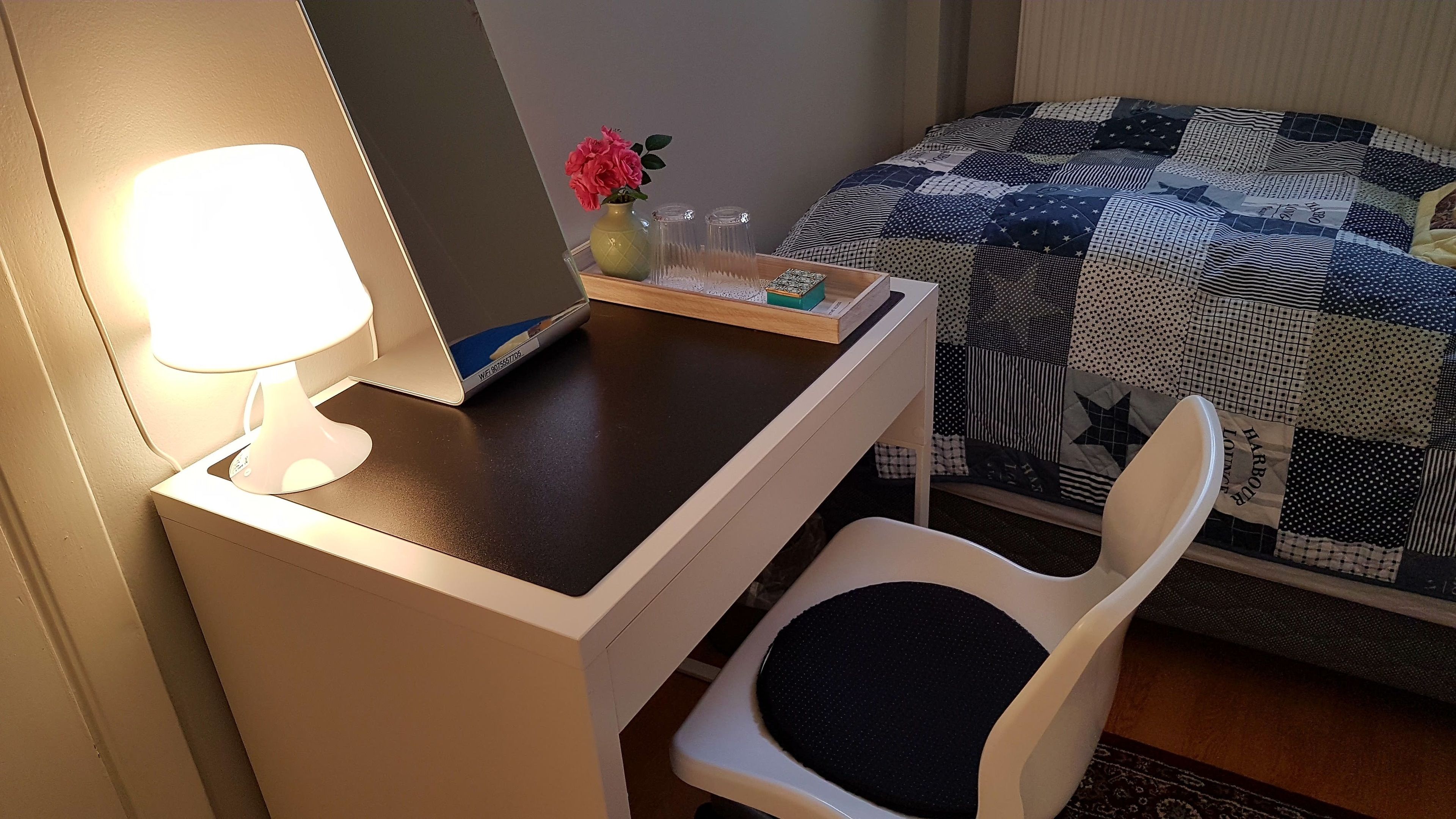 A hotel room with a desk, a chair, a bed with a patchwork quilt, a vase with flowers, and a lamp.