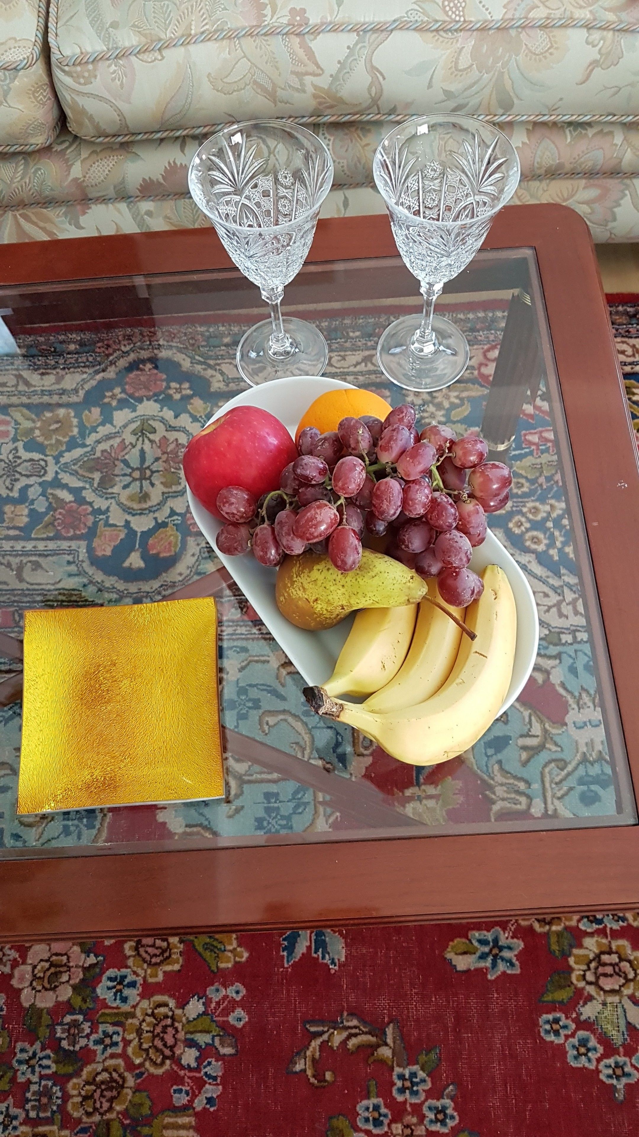 A glass coffee table with a fruit plate, two wine glasses, and a gold-colored object.