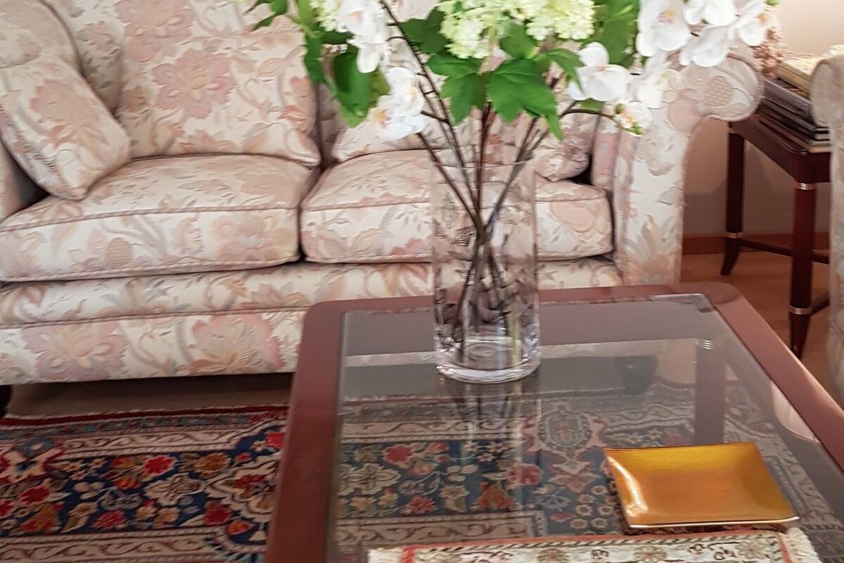 A living room with a floral sofa, a glass coffee table with a vase of flowers, and a patterned rug.