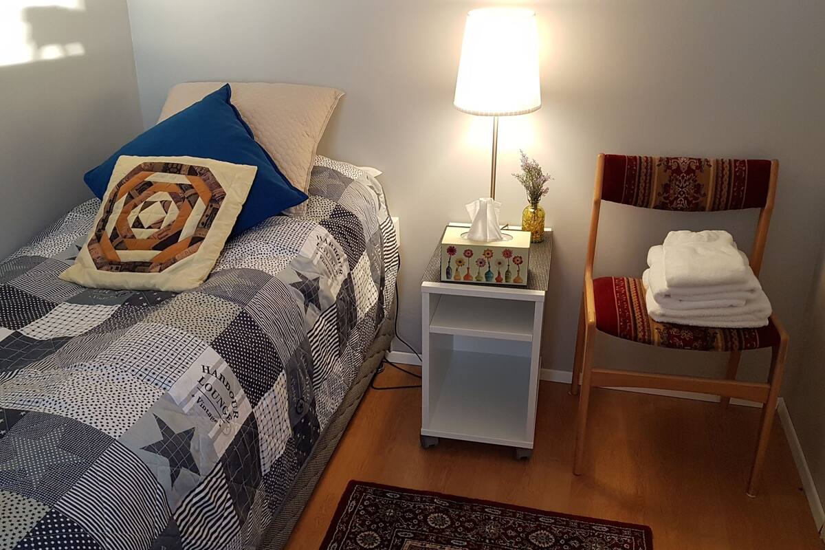 A neatly made bed with a patterned bedspread, a bedside lamp, a small table with a tissue box, a chair with a towel, and a rug on the floor.