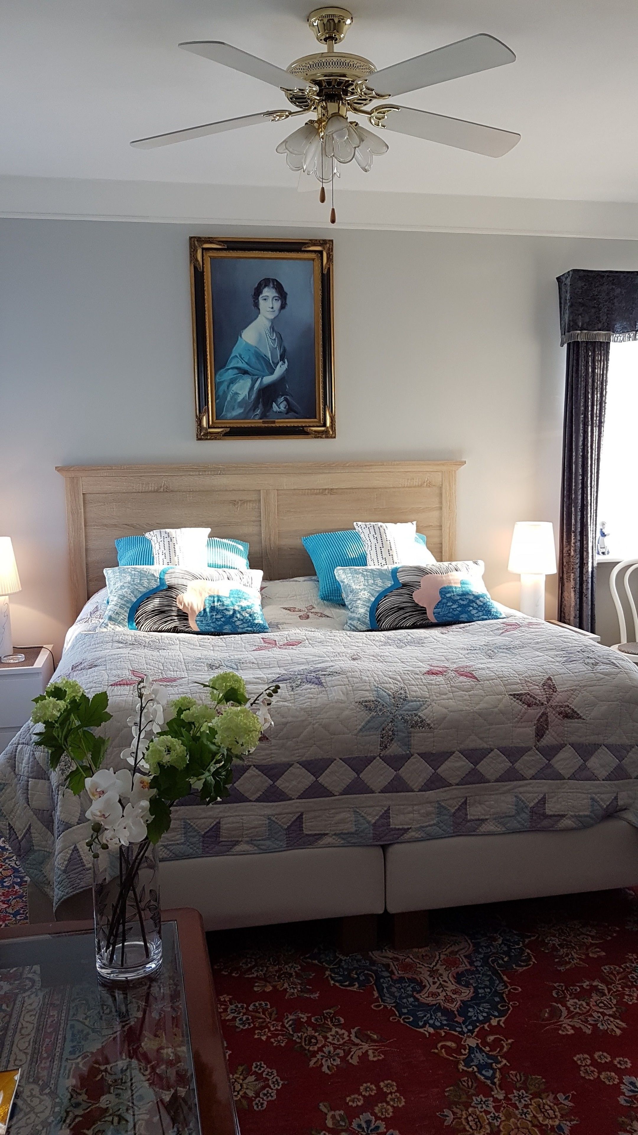 Un dormitorio con una cama grande, un ventilador de techo, un retrato enmarcado y un florero con flores.