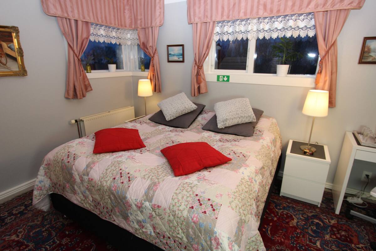 A bedroom with a floral bedspread, two pillows, a nightstand, and a window with curtains.