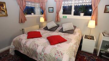 A bedroom with a floral bedspread, two pillows, a nightstand, and a window with curtains.