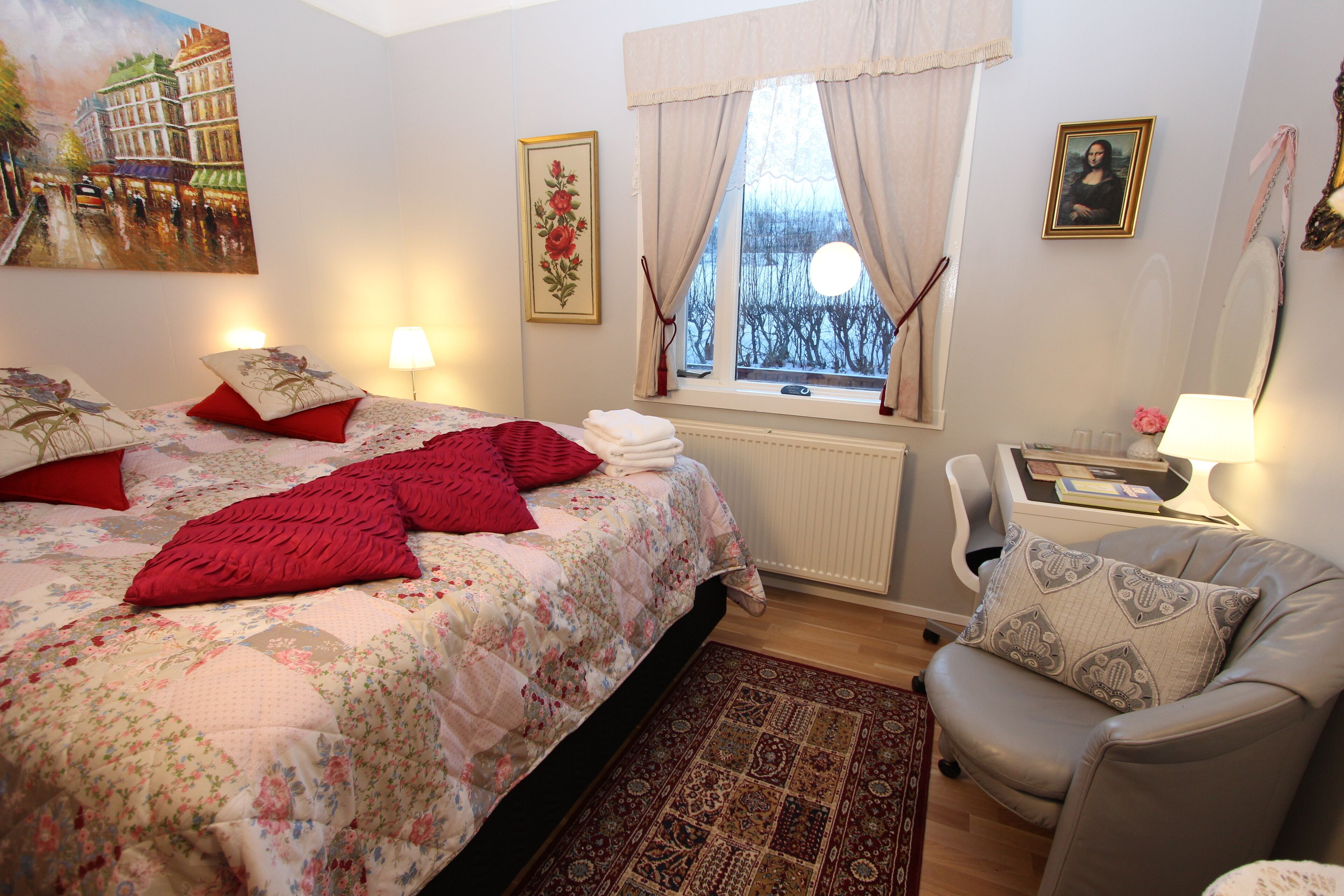 A well-lit bedroom with a bed, a chair, a window with curtains, and a painting on the wall.