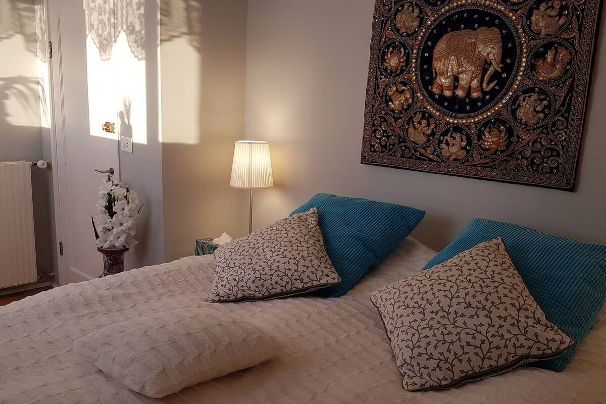 A bedroom with a bed, two pillows, a nightstand, a lamp, a wall decoration, and a window with curtains.