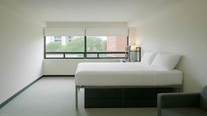 A bedroom with a bed, a nightstand, a lamp, a window, and a view of buildings outside. - Parkside Student Residence (Toronto)