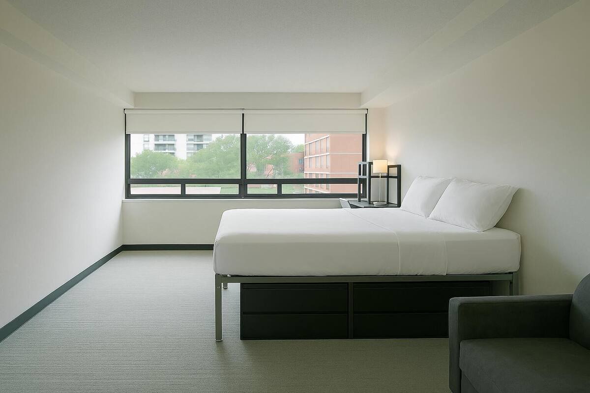 Un dormitorio con cama, mesita de noche, lámpara, ventana y vistas a edificios.
