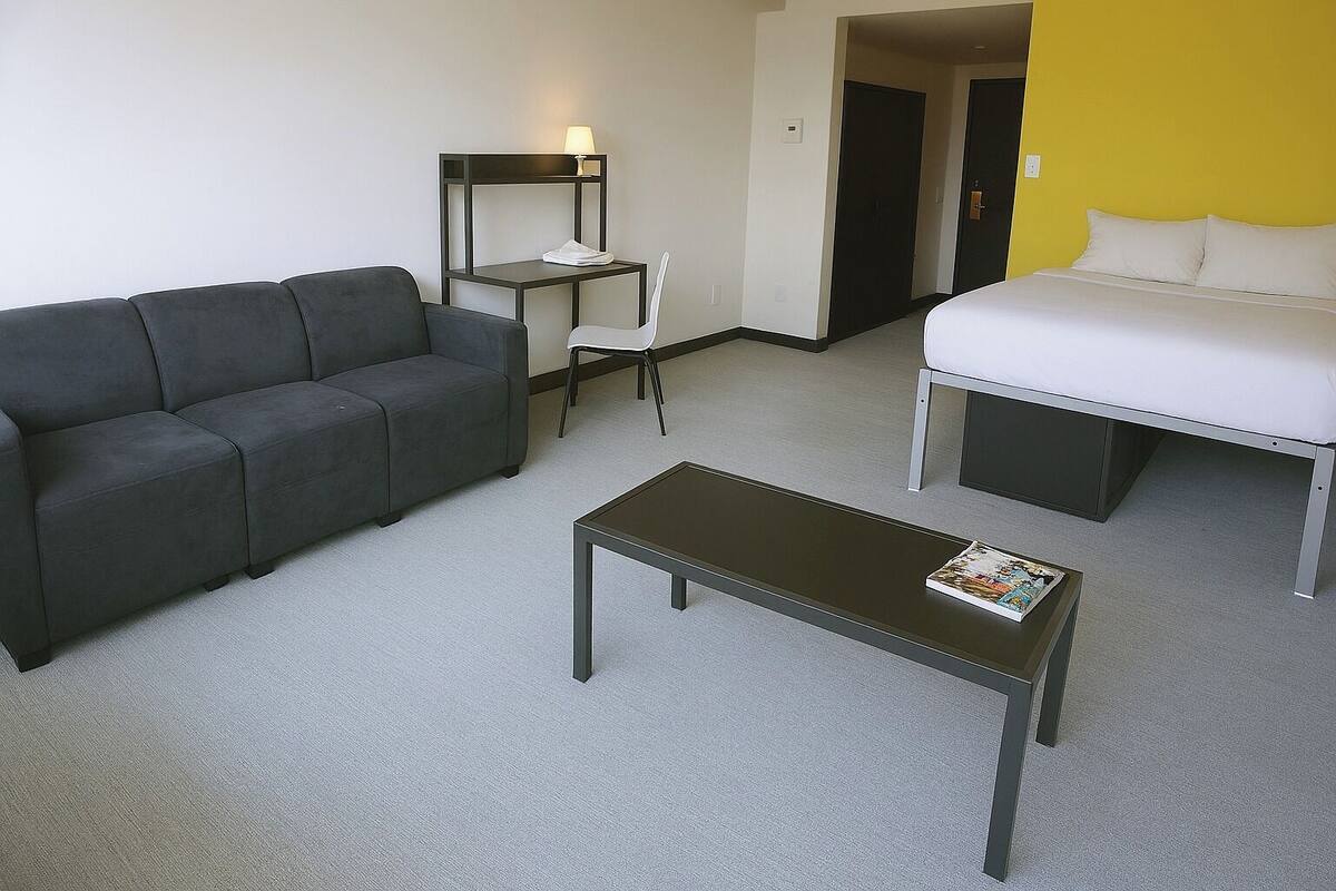 Una habitación con un sofá gris, una mesita con una revista, una silla blanca y una cama con ropa de cama blanca.