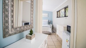 A hotel room with a large ornate mirror, a white dresser, a bed with a white headboard, a nightstand with a lamp, and a TV on a cabinet. - Little Rose Guesthouse & Self-Catering (Gqeberha (Port Elizabeth))
