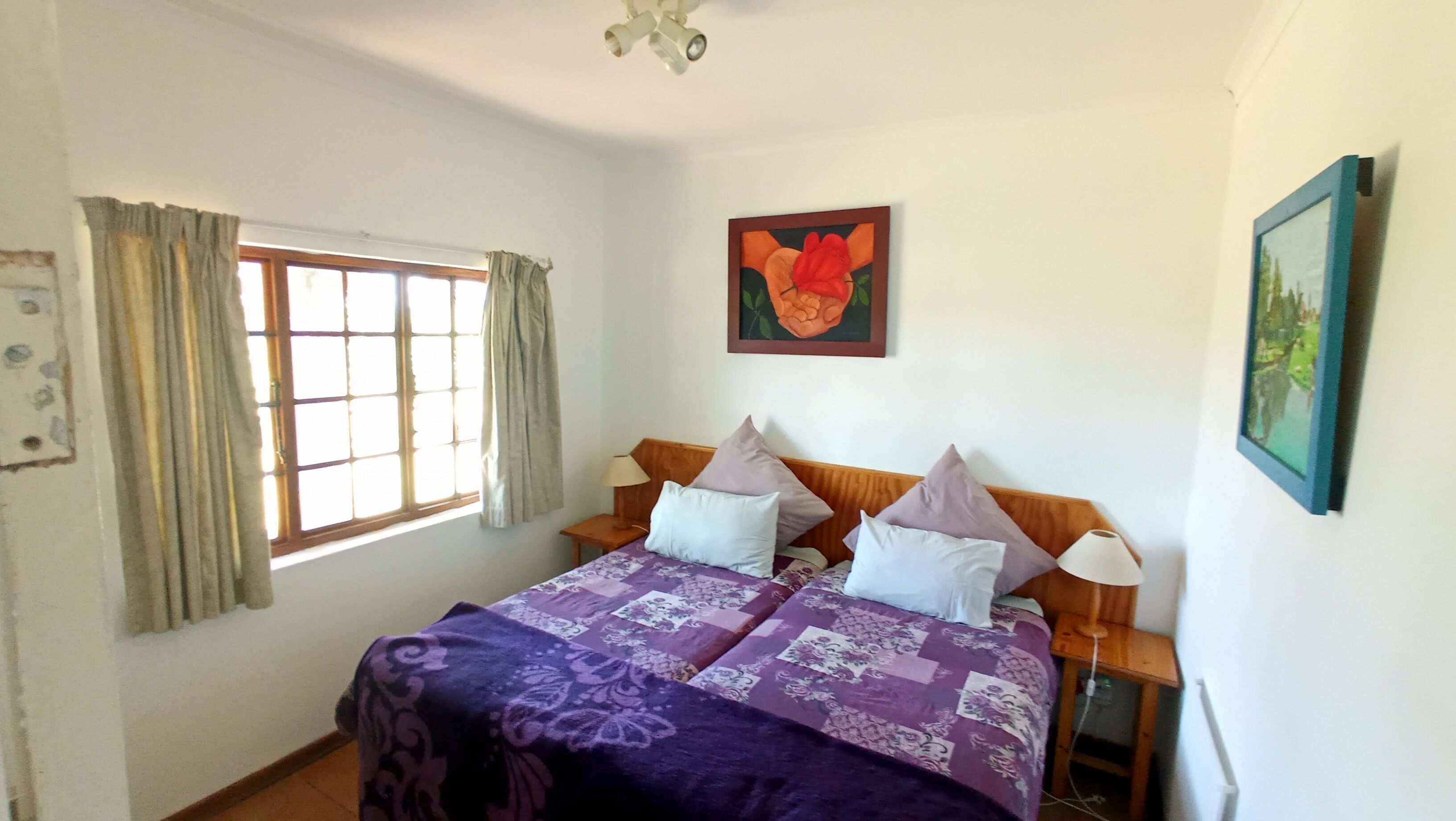 A bedroom with a bed, two bedside tables, a window with curtains, a painting on the wall, and a lamp.