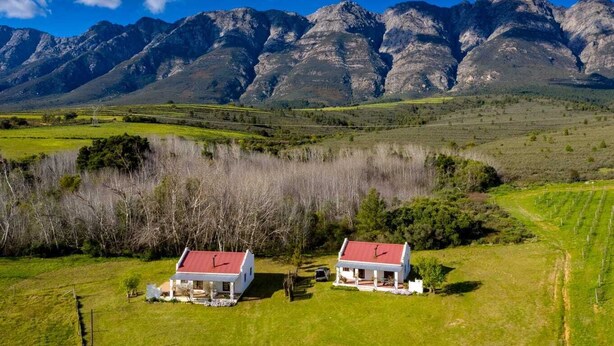 A rural landscape with two white houses with red roofs, surrounded by green fields and mountains in the background. - Fraaigelegen Farm (Tulbagh)