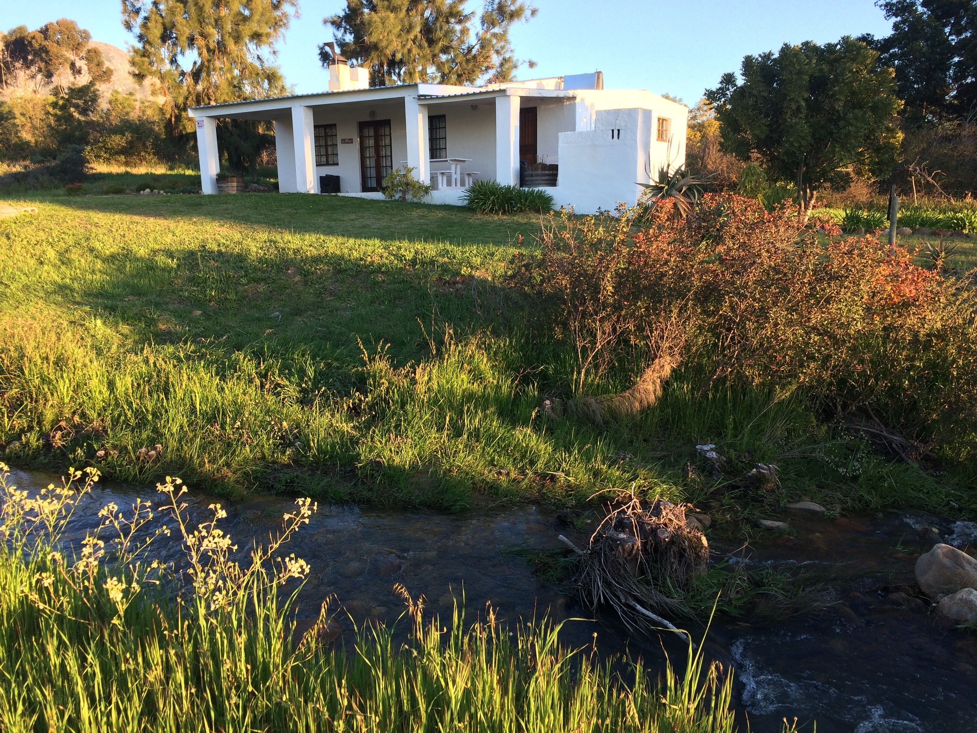 A white single-story house with a porch, surrounded by a grassy field and a small stream.
