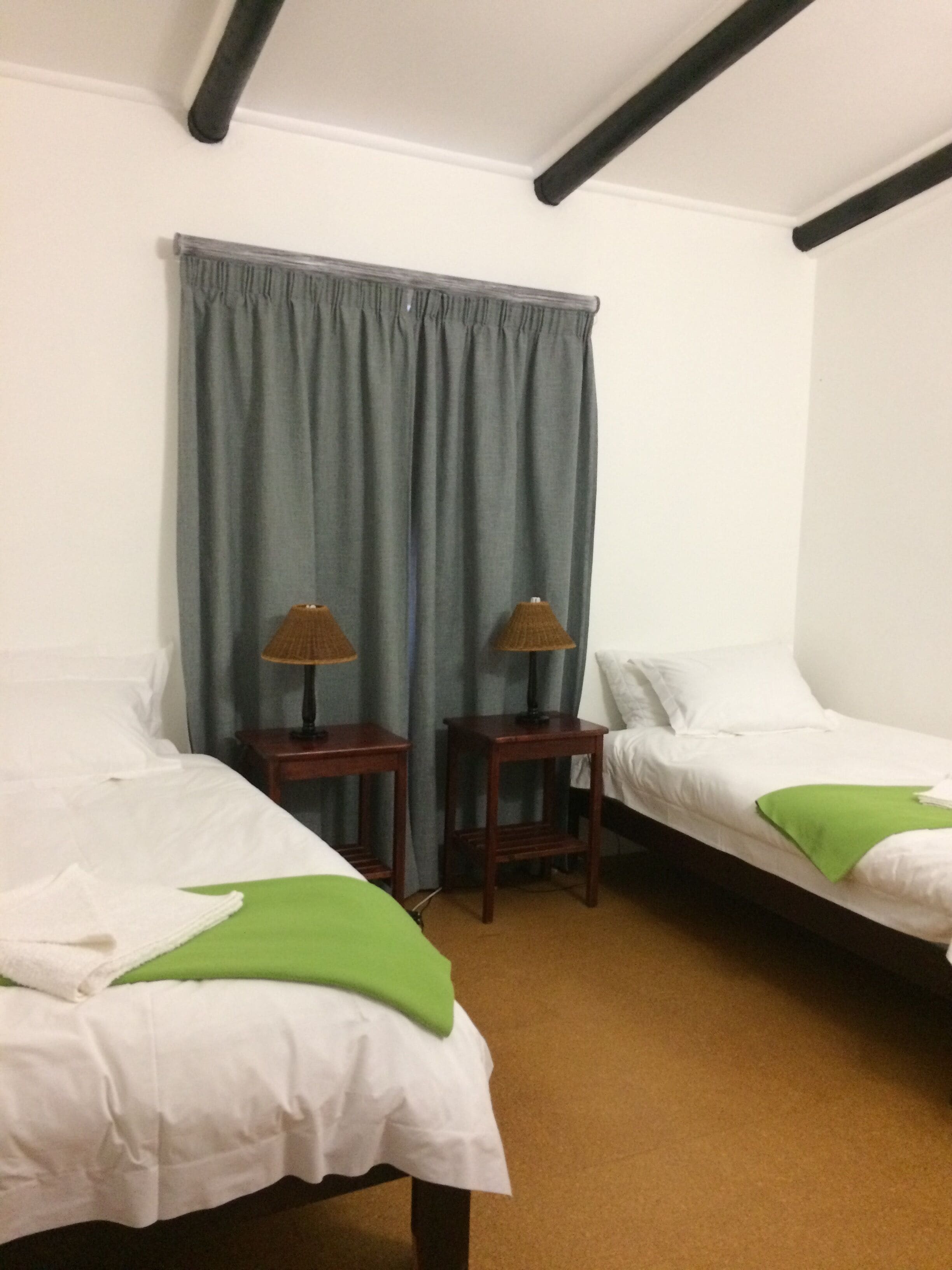 A hotel room with two single beds, a large window with blue curtains, and two bedside tables with lamps.