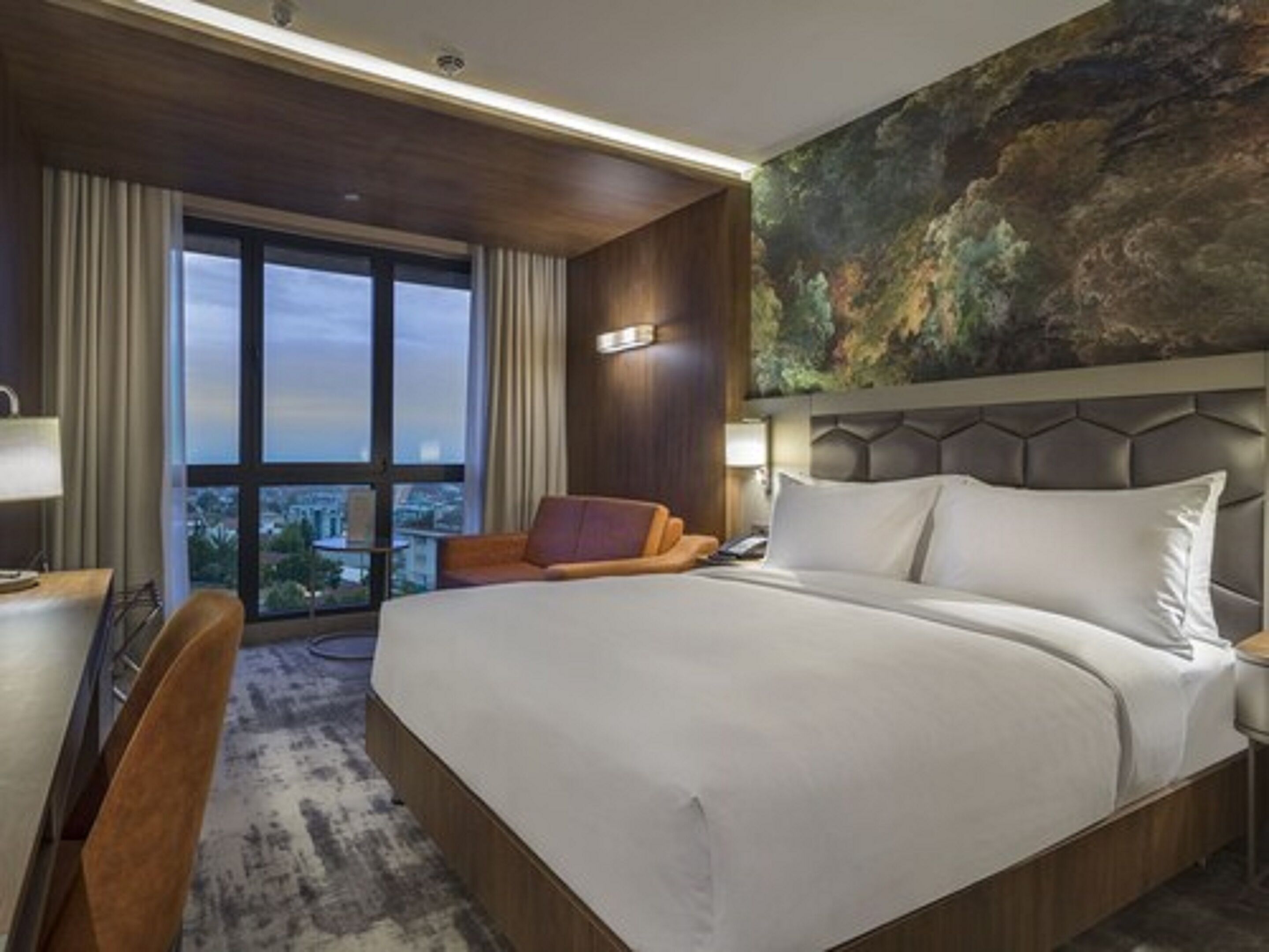 Une chambre d’hôtel avec un grand lit, un bureau, une chaise et une vue sur la ville grâce à une grande fenêtre.