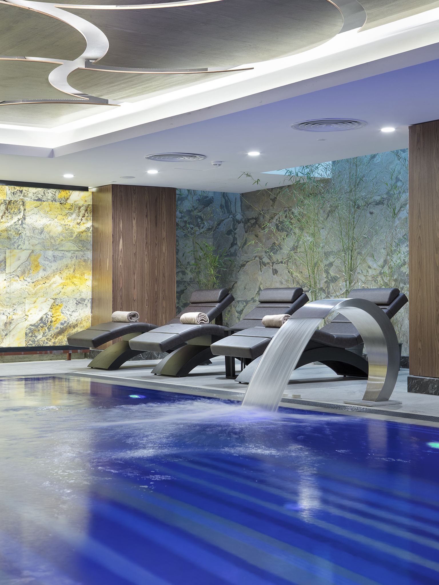 A modern indoor pool area with lounge chairs and a waterfall feature.