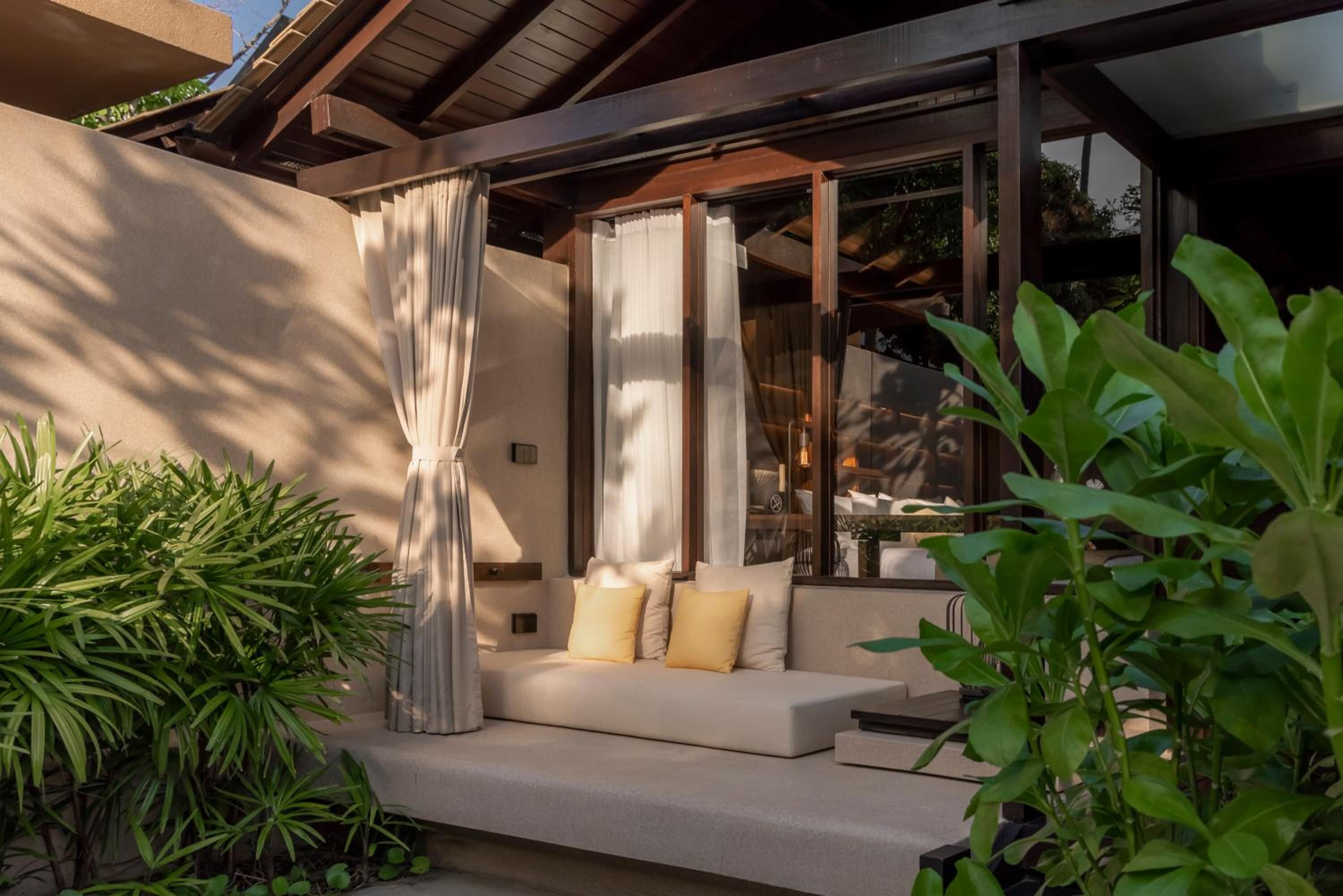 A modern outdoor seating area with a sofa, cushions, and curtains, surrounded by greenery.