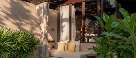 A modern outdoor seating area with a sofa, cushions, and curtains, surrounded by greenery.