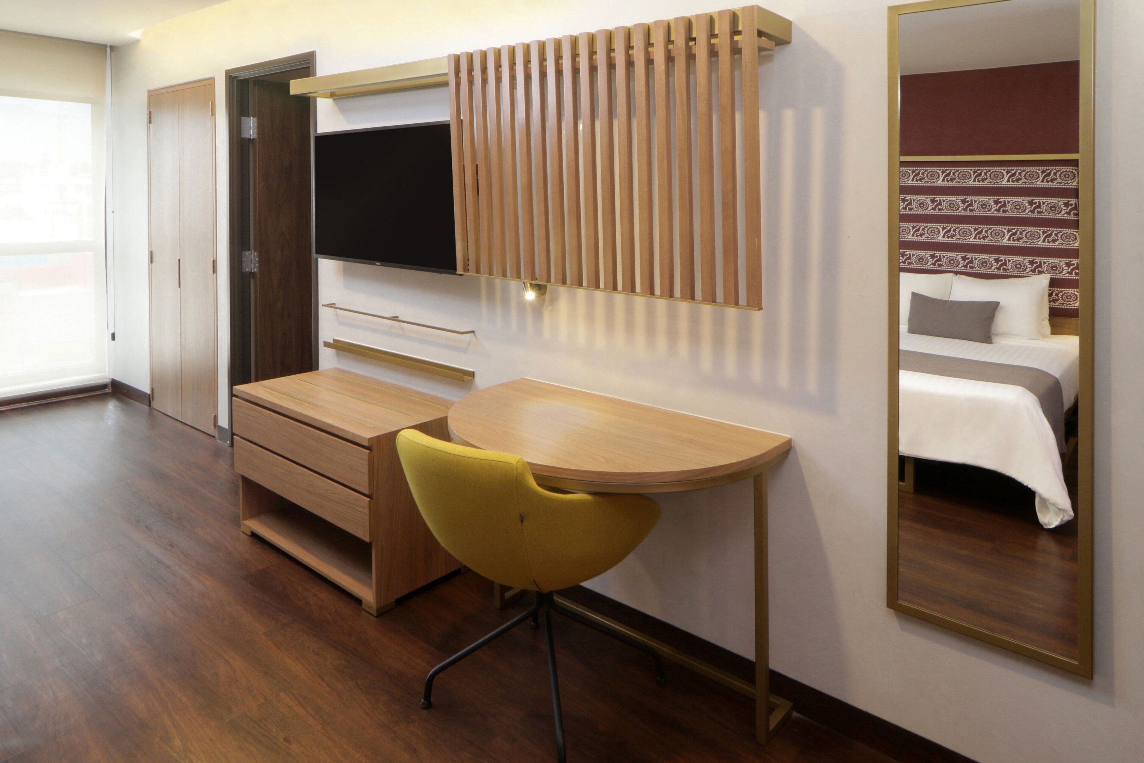 Habitación de hotel con escritorio de madera, una silla amarilla, un televisor montado en la pared y una cama con cabecera estampada.