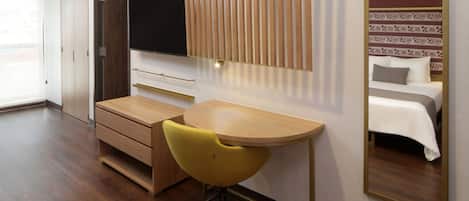 Habitación de hotel con escritorio de madera, una silla amarilla, un televisor montado en la pared y una cama con cabecera estampada.