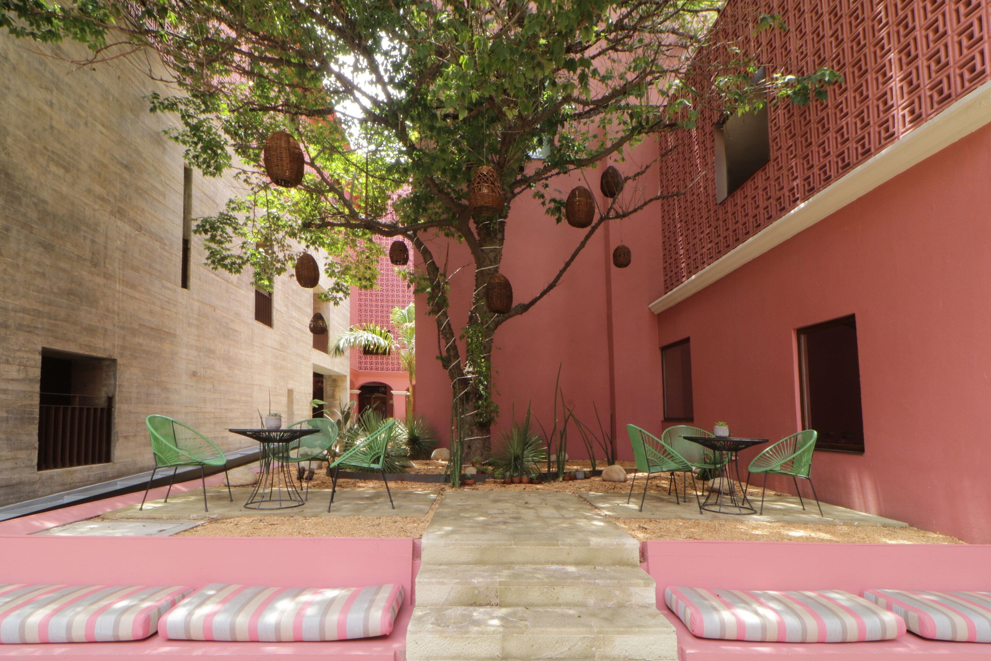 Une cour avec un arbre, des paniers en osier, des chaises vertes et un mur rose.