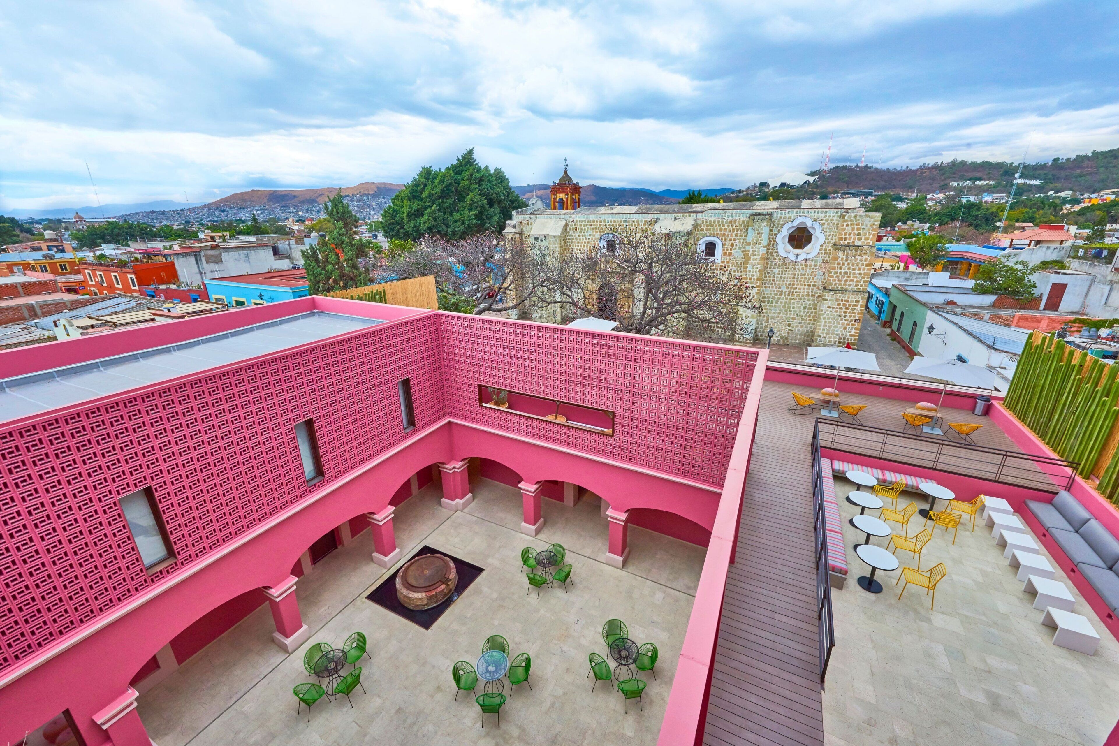 Terraço no telhado com uma parede rosa estampada, assentos ao ar livre em tons de verde e amarelo, e vista para um edifício histórico ao fundo.