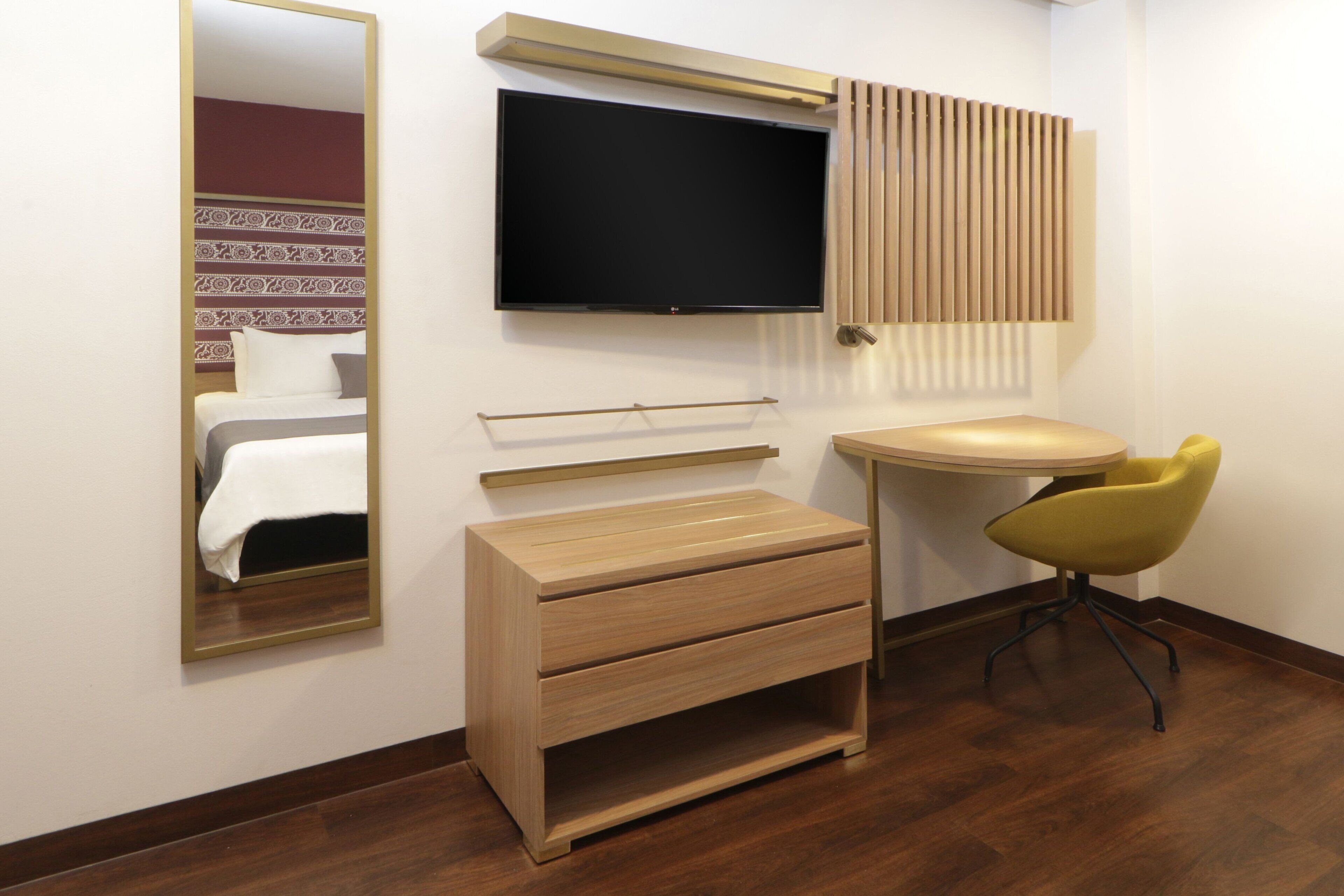 Quarto de hotel com cama, televisão presa à parede, cómoda de madeira, mesa redonda e uma cadeira amarela.