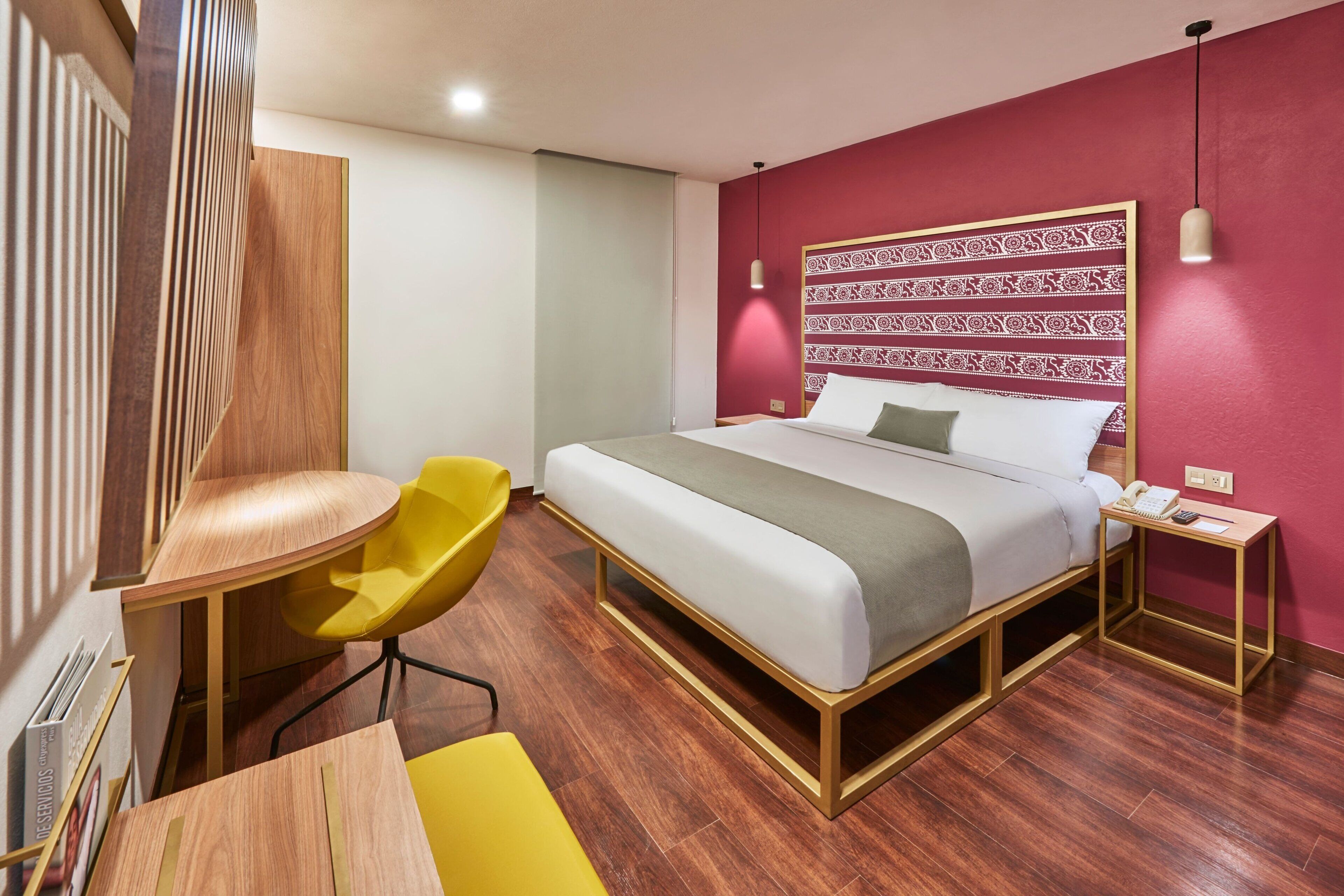 A modern hotel room with a wooden bed frame, a desk, a yellow chair, and a patterned headboard.