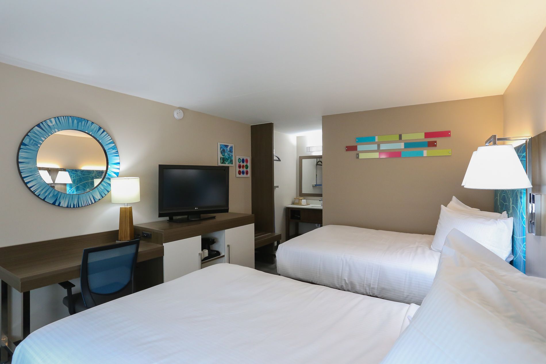 A hotel room with two beds, a desk with a computer, a TV, a mirror, and a bathroom visible through an open door.