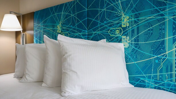 Habitación de hotel con cama, almohadas blancas y una pared con diseño de placa de circuito azul.