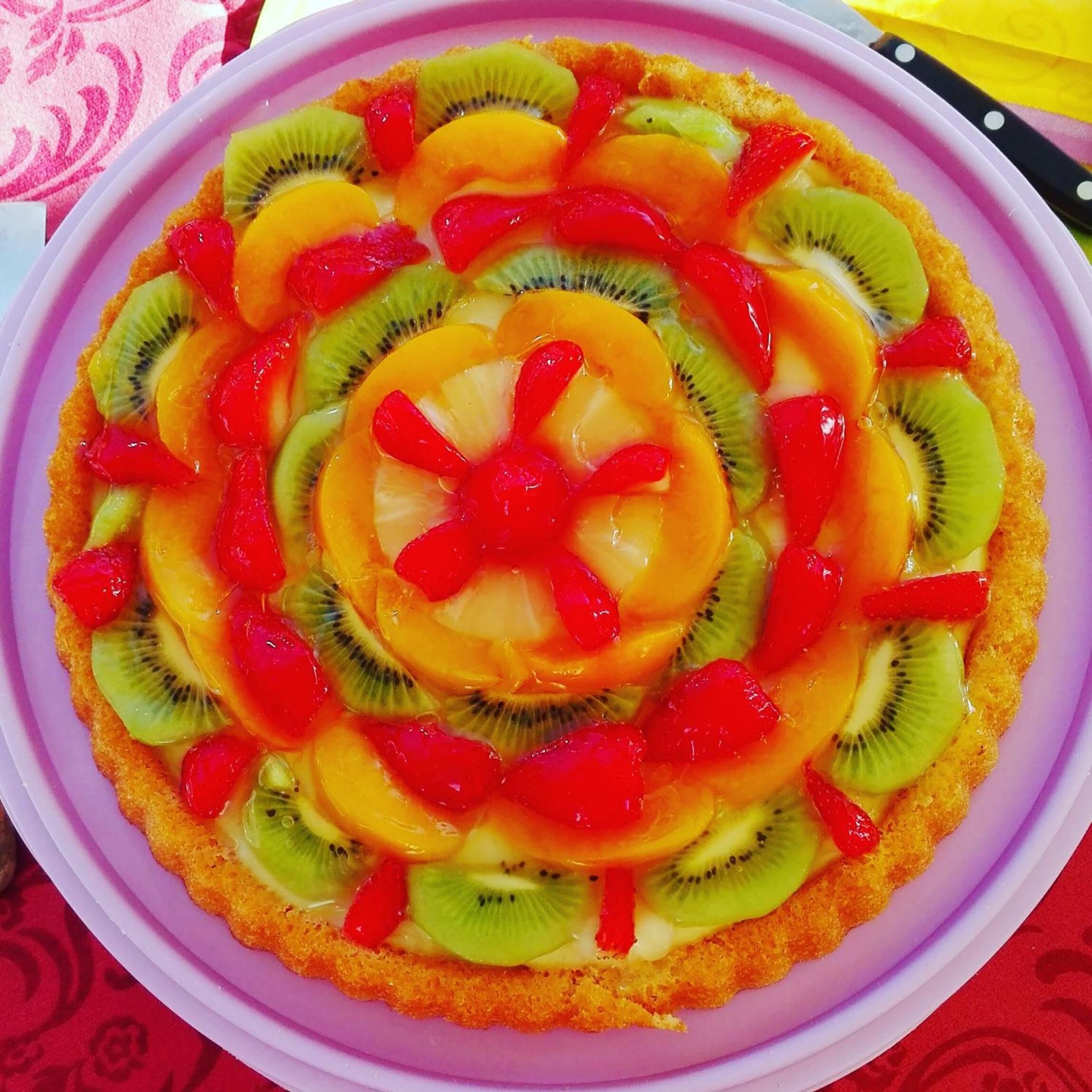 Une tarte aux fruits garnie de tranches de kiwi, d’orange et de fruits rouges, présentée sur une assiette rose.